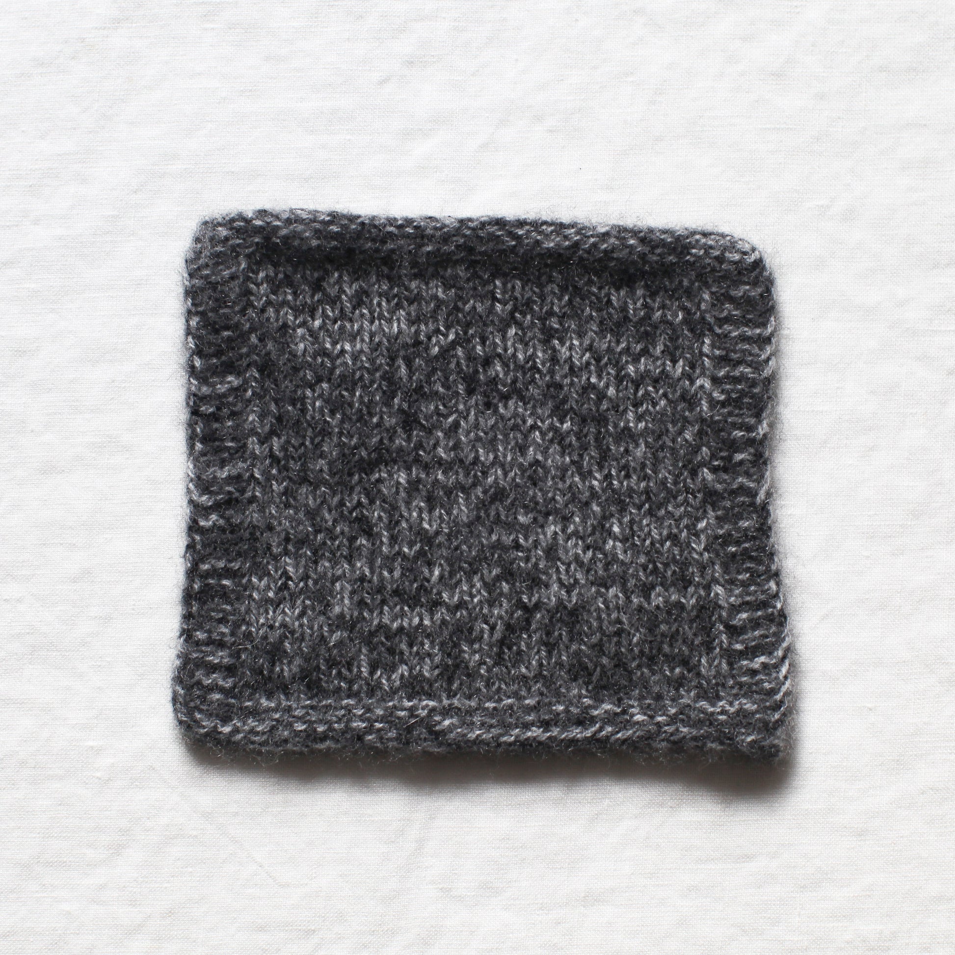 A knitted square from light and dark grey yarns to create a soft marl. The background is a white linen table cloth.