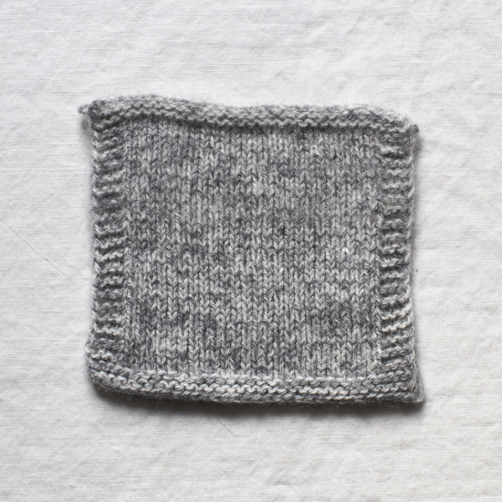 A knitted square made from white and light grey yarns knitted together to create a marl. The background is a white linen tablecloth.