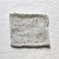 White and a black flecked light grey yarn are knitted into a small sample square to create a gentle marl. The background is a white linen table cloth.