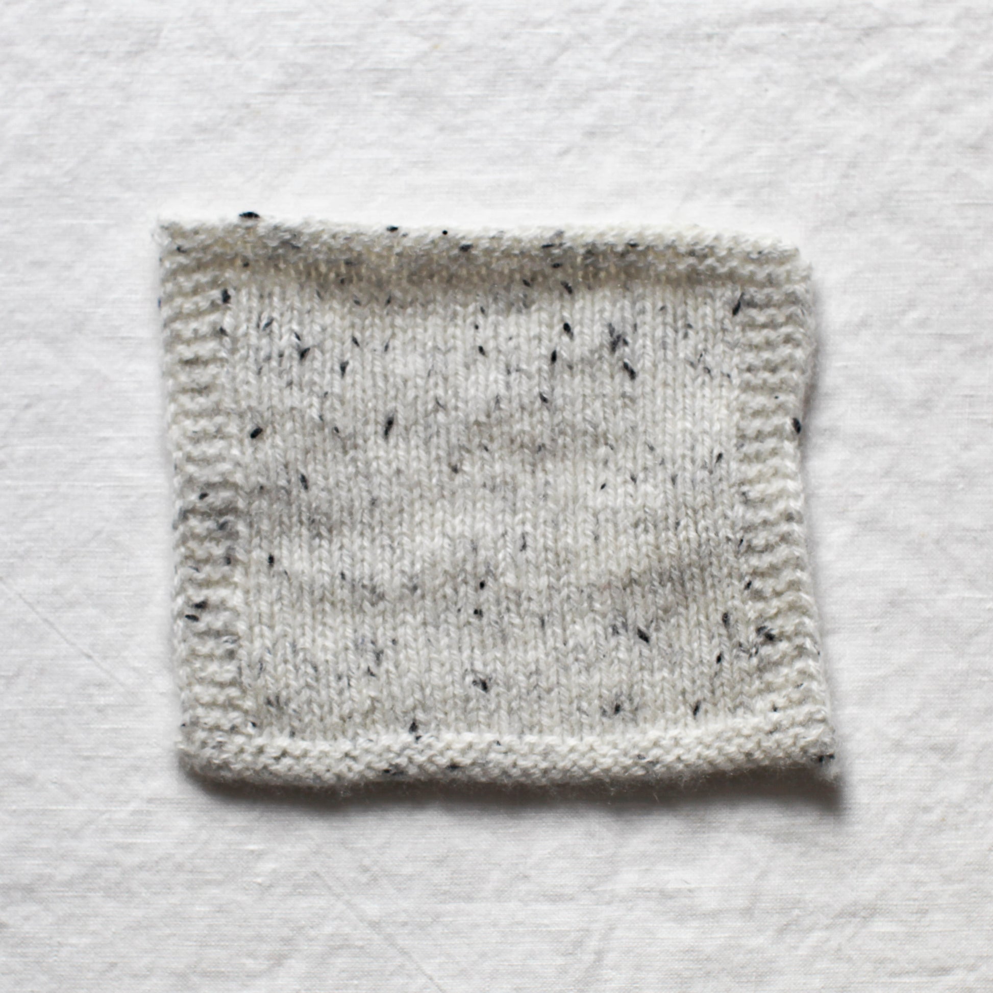 White and a black flecked light grey yarn are knitted into a small sample square to create a gentle marl. The background is a white linen table cloth.